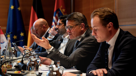 Ministerpräsident Rhein, Staatssekretär Kuhn und weitere Mitglieder der Landesregierung sitzen am Tisch während der Gespräche des Regierungsgipfels. Ministerpräsident Rhein spricht in ein Mikrofon.