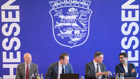 Drei Männer sitzen an einem Tisch im Rahmen einer Pressekonferenz. Im Hintergrund das neue Logo.
