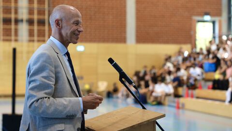 Herr Schwarz steht an einem Rednerpult in einer Turnhalle - im Hintergrund sieht man Schüler und Lehrer