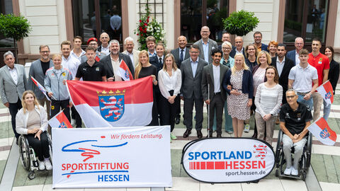 Ministerpräsident Rhein und Sportministerin Stolz stehen inmitten einer Gruppe Sportler, die teilweise Hessen-Flaggen in Händen halten.
