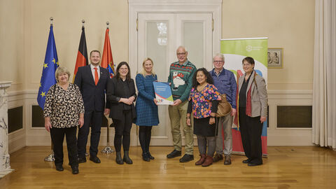 Mehrere Personen stehen vor 3 Flaggen und lächeln in Kamera. Eine Frau überreicht einem Mann eine Auszeichnung.