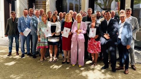 Dr. Sinemus steht in einer Reihe mit mehreren festlich gekleideten Menschen und hält einen Förderbescheid in den Händen.