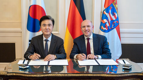 Zwei Männer sitzen am Schreibtisch. Im Hintergrund sind die koreanische, die deutsche und die hessische Flagge zu sehen.