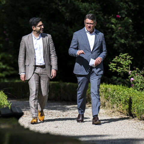 Ministerpräsident Boris Rhein und der stellvertretende Ministerpräsident Kaweh Mansoori laufen in Anzügen nebeneinander einen Weg durch einen grünen Park entlang.